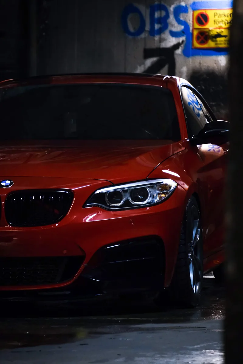 An orange BMW 2 Series coupé in a dim concrete garage, front quarter lit by a strip light.